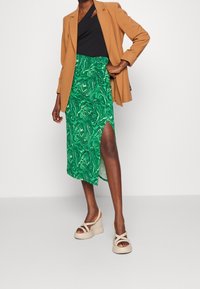 Vihreä marmoroitu hame, jossa on korkea halkio, yhdistettynä mustaan toppiin ja ruskeaan bleiseriin. Asun täydentävät beiget korolliset sandaalit.