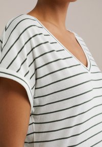 Vit t-shirt med v-ringning och mörkgröna horisontella ränder, korta ärmar, mjuk bomullstextur och kontrasterande kant längs halsringningen.