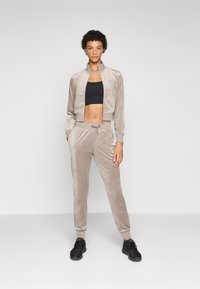 Velvet loungewear set in light beige, featuring a cropped zip-up jacket and tapered joggers with a drawstring. Black sports bra underneath.