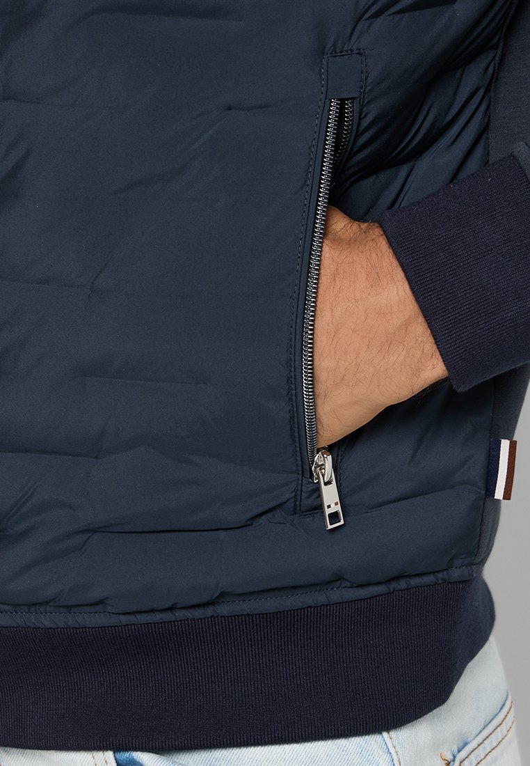 Giacca isolante blu navy con tasca con zip, polsini e orlo a coste, e una texture trapuntata orizzontale. Accentuata da un piccolo logo.