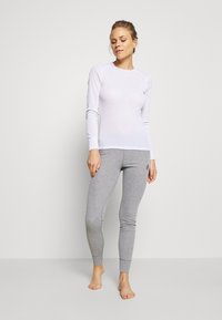 Weißes Langarmoberteil mit Rundhalsausschnitt und gerippten Bündchen, kombiniert mit enganliegenden grauen Leggings, die ein kleines Logo am linken Oberschenkel haben.
