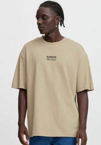 Beige cotton t-shirt with short sleeves and relaxed fit. Features black text "Actions Decisions" printed on the front.