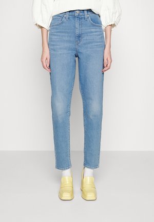 Personne portant un jean droit taille haute bleu clair, associé à des chaussettes blanches et des mocassins jaune pastel, debout devant un fond blanc.