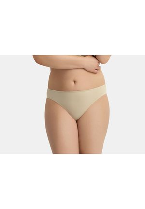 Donna che indossa biancheria intima stile bikini beige con le braccia incrociate sul torso nudo su uno sfondo semplice grigio chiaro.