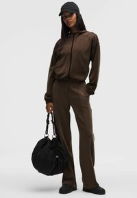 Cazadora con cremallera marrón y pantalones a juego hechos de un tejido suave. Gorra negra y un bolso grande negro con un diseño fruncido. Calzado resistente.