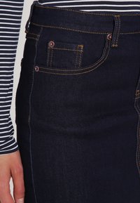 Jupe en denim foncé avec des coutures jaunes, dotée de poches frontales et de boutons cuivrés. Portée avec un haut à rayures à manches longues.