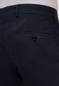 Marineblaue maßgeschneiderte Hose aus strukturiertem Stoff, mit einer Gesäßtasche und einem schwarzen Knopf sowie subtilen Karomuster-Details.