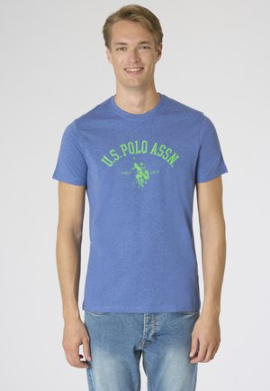 Giovane uomo sorridente, indossa una t-shirt blu U.S. Polo Assn. con logo verde e jeans azzurri, in piedi davanti a uno sfondo semplice.