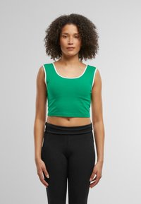Grön kortad linne med vit kant, tillverkad av stretchigt material. Modellen har kombinerat den med högmidjade svarta leggings och står framför en ljus bakgrund.