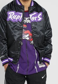 Chaqueta bomber negra de satén con gráfico púrpura de "Raptors", puños a rayas púrpura y blanca, sobre una camiseta de baloncesto púrpura con un dinosaurio rojo.