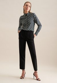 Camicia a maniche lunghe a fantasia in blu navy con un design geometrico bianco, abbinata a pantaloni neri su misura e sandali neri con cinturino. Sfondo semplice.