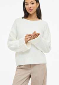 Biały, miękki sweter dziany z szerokim dekoltem, ściągaczowymi mankietami i luźnym krojem, zestawiony z beżowymi zwężanymi spodniami.