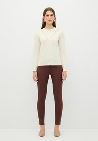 Cremefarbenes Langarmshirt mit V-Ausschnitt und roten Nähten, kombiniert mit braunen Leggings und nude-farbenen High Heels, was eine taillierte Silhouette schafft.