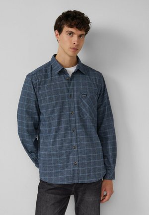 Junger Mann mit lockigem Haar, der ein blau kariertes Hemd über einem weißen T-Shirt trägt, steht vor einer schlichten hellgrauen Wand.