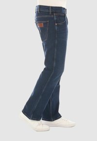 Jeans de denim azul oscuro con piernas rectas, diseño de cinco bolsillos y detalle de cuero marrón en el bolsillo trasero. Llevados con zapatillas blancas.