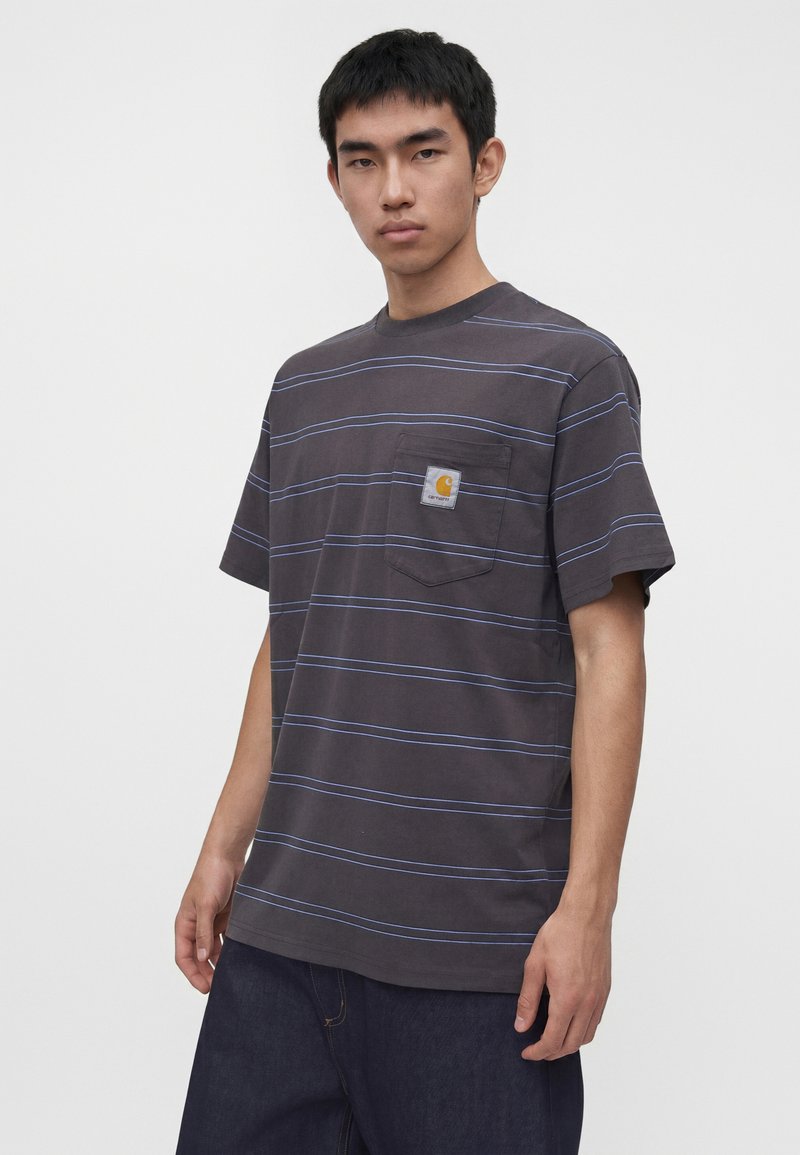 Giovane uomo che indossa una T-shirt a righe grigio scuro con taschino sul petto e toppa con logo giallo, in piedi su uno sfondo bianco semplice.