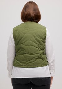 Olivgrüne, gesteppten Weste mit einem wellenförmigen Muster, getragen über einem weißen Langarmshirt. Sie verfügt über einen runden Halsausschnitt und einen Saum, der auf der Taille endet.