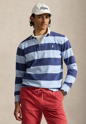 Jeune homme portant une casquette blanche Ralph, un maillot de rugby rayé bleu et bleu clair, un short rouge à cordon et une montre-bracelet, posant avec une main dans la poche.