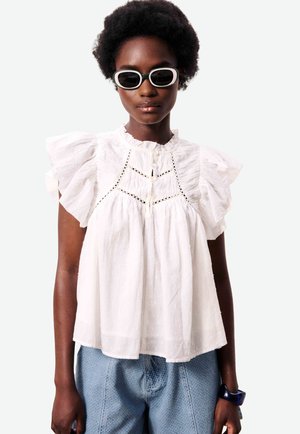 Vrouw draagt een witte blouse met ruches en kanten details, blauwe textuurige broek, zwart-witte zonnebril en een donkere armband aan haar rechterpols.