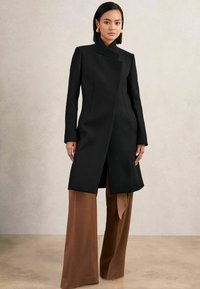 Cappotto in misto lana nero con colletto alto, fronte asimmetrico e silhouette sagomata, abbinato a pantaloni marroni a gamba larga.