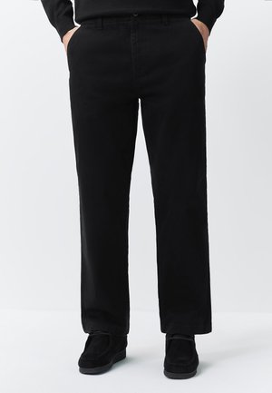 Osoba oblečená v čiernych nohaviciach s rovnými nohavicami, s rukami v kapse a čiernymi topánkami, stojaca pred jednoduchým svetlým pozadím.