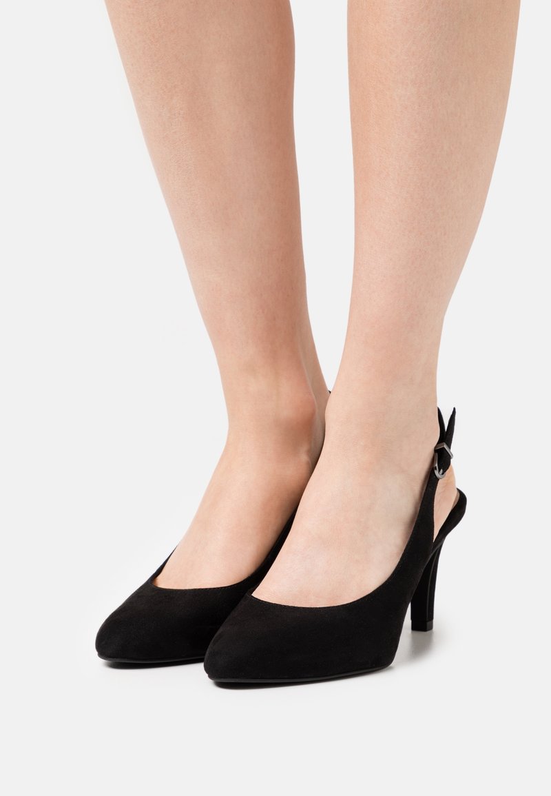 Paire d'escarpins slingback en daim noir avec bouts pointus et brides à la cheville, portés pieds nus sur un fond uni.