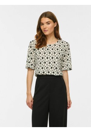 Femme aux cheveux bruns portant une blouse blanche à motifs floraux noirs et un pantalon noir taille haute, debout devant un fond clair uni.