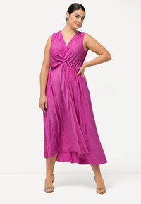 Fuchsiové plisované šaty s výstřihem do V a bez rukávů. Vyznačují se zkrouceným živůtkem a splývavou sukní. Nosí se s vysokými botami na průhledných popruzích.