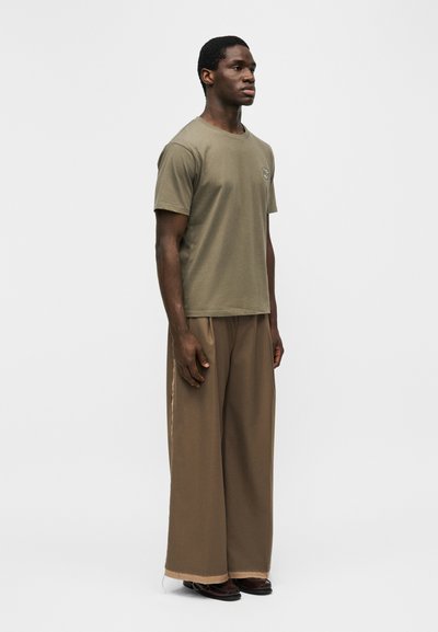 Νεαρός άνδρας που στέκεται στραμμένος προς τα δεξιά, φοράει πουκάμισο λαδί και φαρδιά καφέ παντελόνια με σκούρα παπούτσια, ουδέτερο γκρι φόντο.