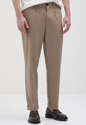 Männliches Model in beigefarbenen maßgeschneiderten Hosen mit Umschlägen und schwarzen Lederloafers, stehend vor einem schlichten weißen Hintergrund.
