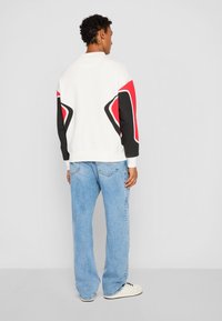 Hvit sweatshirt med svarte og røde geometriske motiver på ermene, sammen med lyseblå vide jeans og hvite joggesko.