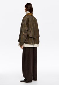 Przesadny plaidowy żakiet w ciemnozielonym, brązowym i złotym odcieniu, z kołnierzem z korduroju i warstwowym krojem, zestawiony z luźnymi brązowymi spodniami.