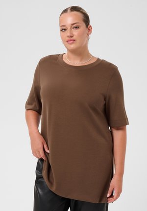 Femme aux cheveux tirés en arrière porte un haut marron à manches courtes et un pantalon noir, accessoirisés de boucles d'oreilles en perles et d'un collier fin.