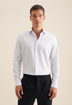 Camicia bianca a fantasia con maniche lunghe, chiusura con bottoni e colletto. Abbinata a pantaloni scuri, che mostrano una vestibilità ordinata.