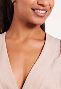 Beige V-hals top met subtiele verticale strepen, toont huidskleur in het nekgebied en een geperforeerd oor met een ronde blauwe edelsteenoorbel.