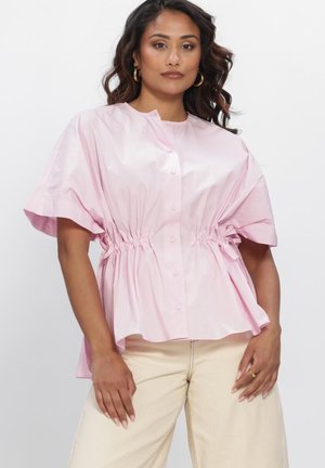 Vrouw met golvend haar draagt een losse, lichtroze blouse met knopen en een tunnelkoord in de taille en beige broek tegen een effen witte achtergrond.