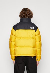 Gul og sort pufferjakke med høj krave, vandret stikninger og The North Face-logo på den øverste ryg.