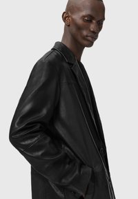 Giacca in pelle nera con colletto stondato, chiusura con singolo bottone e tasche laterali. Texture liscia e silhouette rilassata.