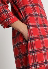 Chemise en flanelle à carreaux rouges avec des rayures noires et jaunes, à manches longues, et une poche latérale. Texture douce avec poignets à boutons et coupe décontractée.