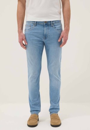 Man die lichtblauwe slim-fit jeans, een wit T-shirt en beige suède loafers draagt, staand tegen een lichte achtergrond.