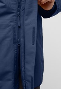 Giacca blu navy con superficie liscia e strutturata. Presenta una cerniera centrale e dettagli in Velcro lungo i lati per una chiusura sicura.