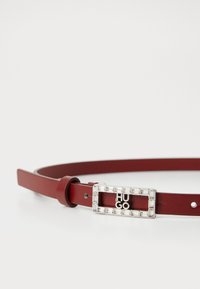 Punainen nahkavyö, jossa on suorakulmainen hope buckle, jossa on teksti "HUGO" ja kirkkaat kivet sen ympärillä, muotoiltu sileällä pinnalla.