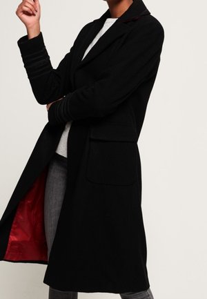 Osoba ubrana w długi czarny płaszcz z czerwonym podszyciem, szare spodnie i jasny sweter, pozująca z jedną ręką skrzyżowaną, a drugą uniesioną.