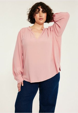 Vrouw met krullend haar die een losse roze blouse met lange mouwen en donkerblauwe broek draagt, poseert met één arm achter haar hoofd opgeheven.