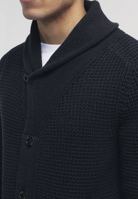 Marinblå stickad kavaj med sjalkrage, med en strukturerad väv och svarta knappar, uppvisad på en neutral bakgrund.