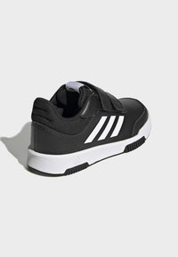 Scarpa da ginnastica nera con una tomaia sintetica liscia, branding a tre strisce bianche, cinturino con chiusura a strappo e una suola in gomma testurizzata.