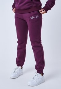 Pantalones de chándal burdeos con puños elásticos, que cuentan con bolsillos laterales y un detalle del logotipo blanco en el bolsillo derecho, combinados con zapatillas blancas.