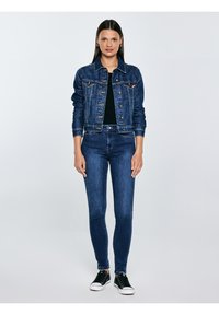 Sieviete stāv, skatoties tieši priekšā, ģērbusies tumši zilā džinsa jaku, saskaņotās džinsās, melnā kreklā un melnos sporta apavos ar baltām zolēm.