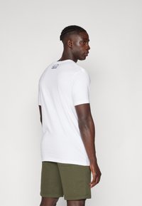 Camiseta blanca de manga corta, de material de algodón, vista trasera con un pequeño logo negro en el cuello. Llevada con pantalones cortos de color verde oliva.