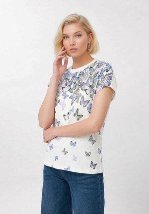 Femme blonde portant un t-shirt blanc avec des motifs de papillons violets et noirs et un jean bleu, posant avec la main près du visage.